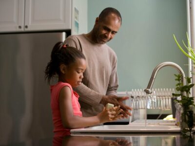 a father and daughter stand together at a sink doing dishes
