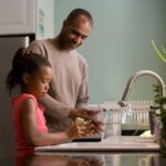 a father and daughter stand together at a sink doing dishes