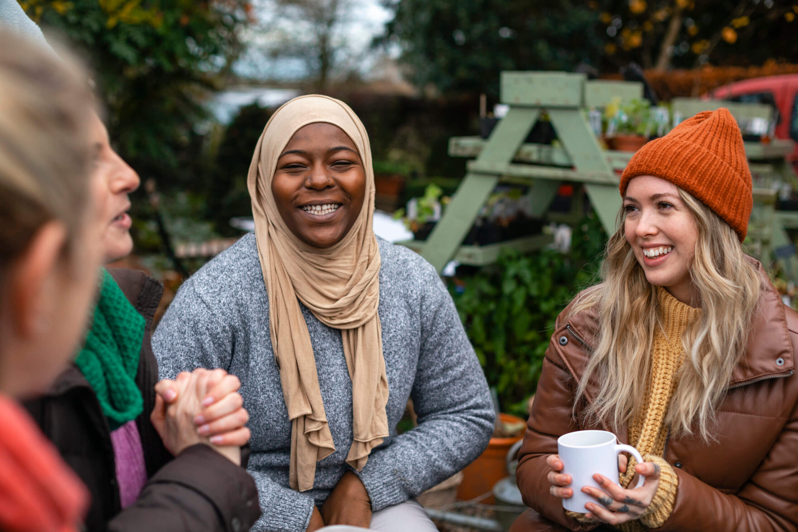 Volunteers sitting outdoors wearing warm casual clothing on a sunny cold winters day. They are resting and having a tea break from working on a community farm, looking after crops and performing other sustainable and environmentally friendly tasks. They are laughing and talking together, drinking hot drinks.